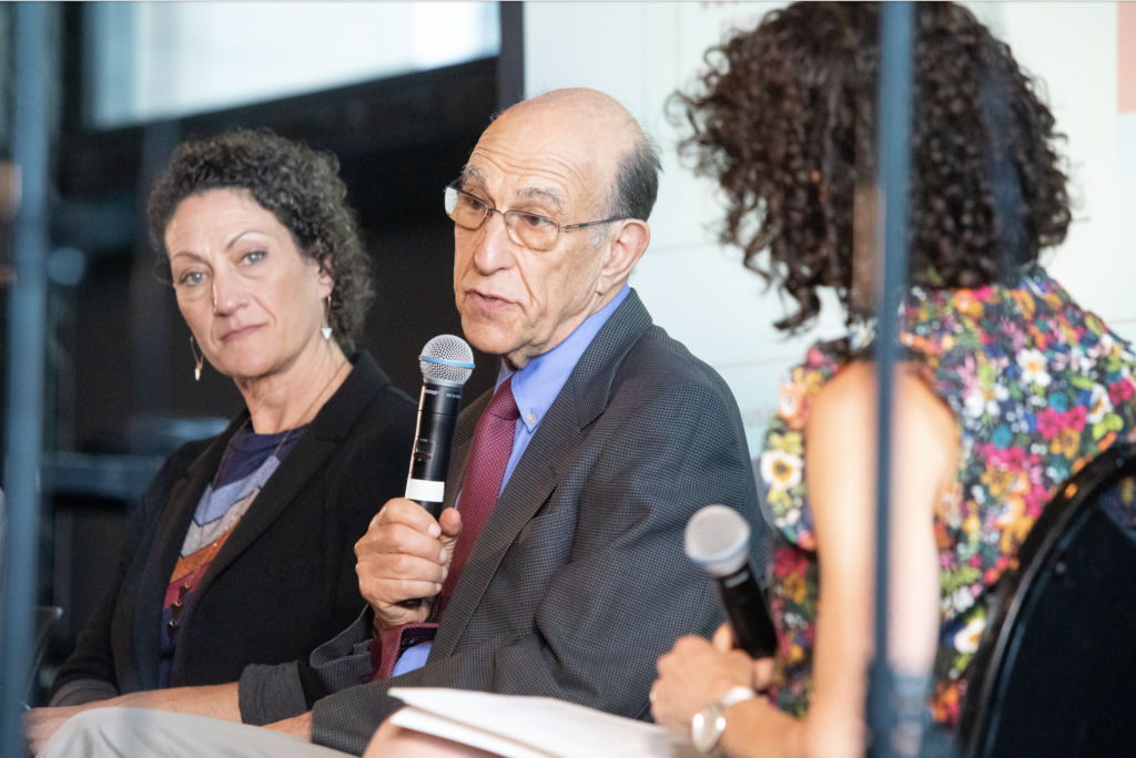 Chicago Housing Commissioner Marisa Novara sits next to Leah and Richard Rothstein. Richard Rothstein speaks into a microphone.