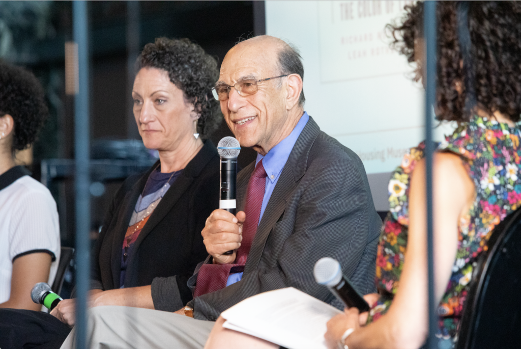 Chicago Housing Commissioner Marisa Novara sits next to Leah and Richard Rothstein. Richard Rothstein speaks into a microphone.