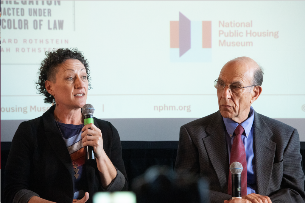 Leah Rothstein speaks into a microphone while sitting in front of a screen with the National Public Housing Museum's logo on it. Richard Rothstein sits next to her.