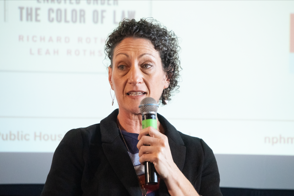 Leah Rothstein speaks into a microphone while sitting in front of a screen with the National Public Housing Museum's logo on it.