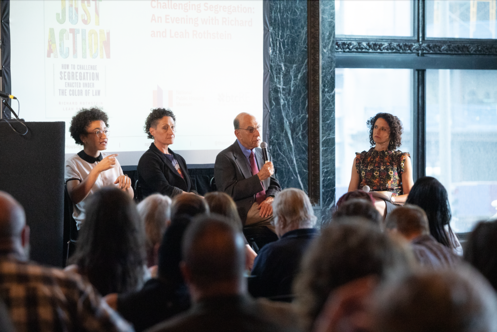 ASL interpreter Makeda Duncan, Leah Rothstein, Richard Rothstein, and Marisa Novara sit in a row in front of a screen. They face a large crowd.