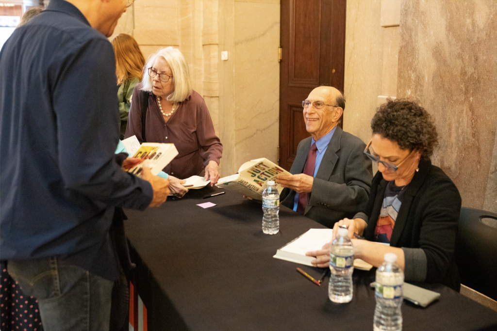 Richard and Leah Rothstein sit at a table and sign books for people standing on the other side of the table.