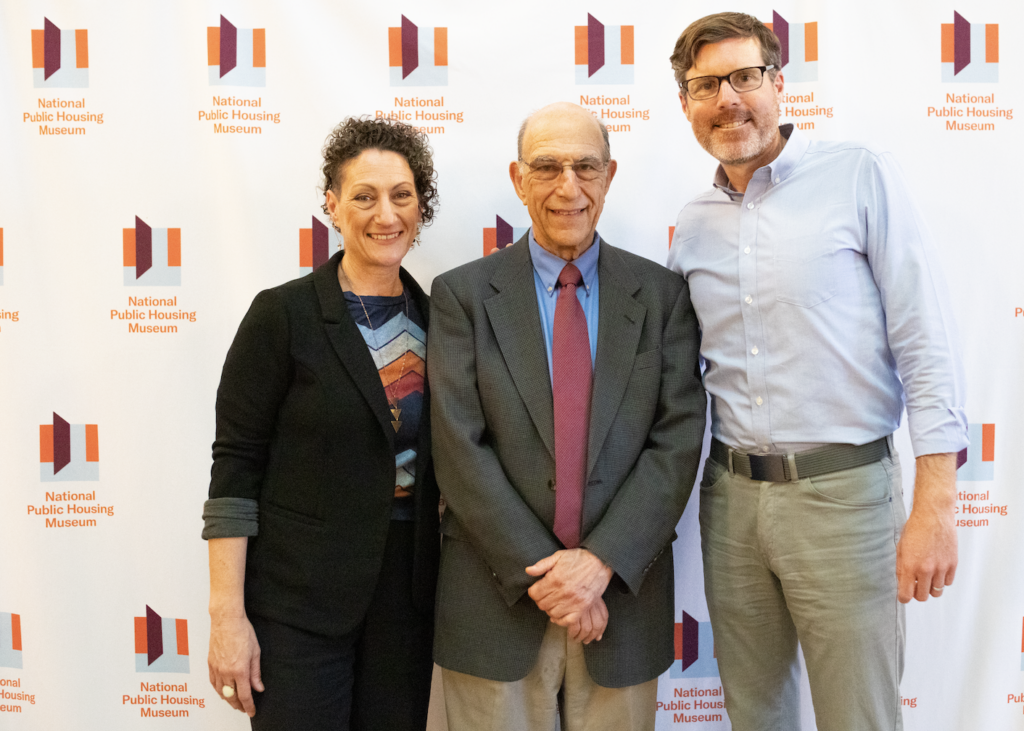 Authors Richard and Leah Rothstein stand next to co-founder of btcRE, M. Ryan Gorman, the sponsor of the event. They all stand in front a a branded white banner with small National Public Housing Museum logos repeated across it.