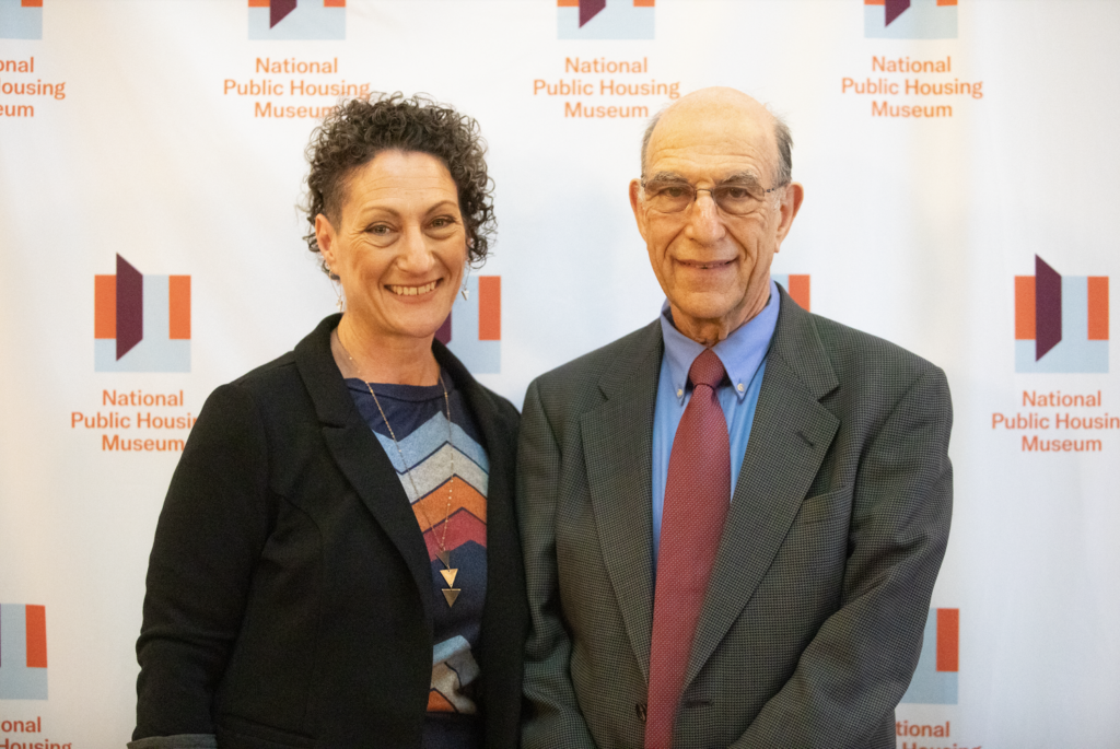 Authors Richard and Leah Rothstein stand in front a a branded white banner with small National Public Housing Museum logos repeated across it.