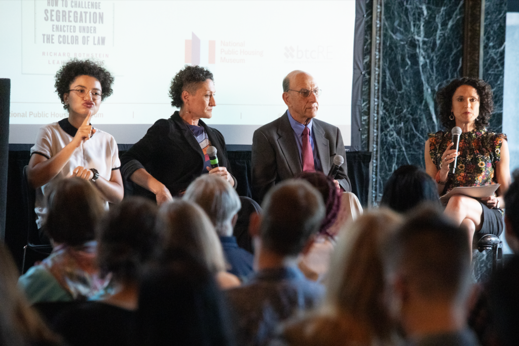 ASL interpreter Makeda Duncan, Leah Rothstein, Richard Rothstein, and Marisa Novara sit in a row in front of a screen. They face a large crowd.