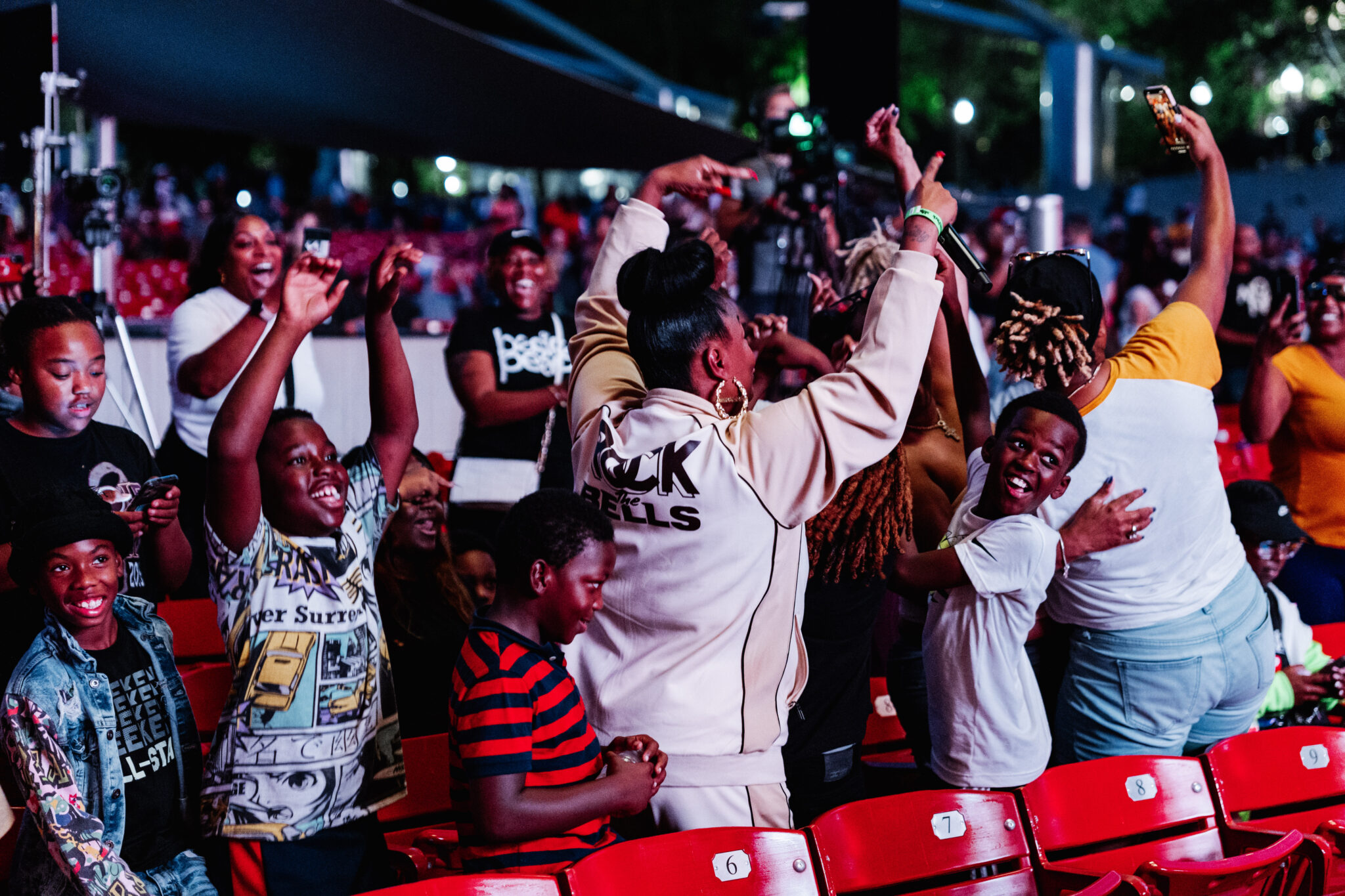 At night in a concert setting, a group of kids dance with a Black woman holding a microphone. The crowd of young people and adults are joyously dancing and enjoying music.