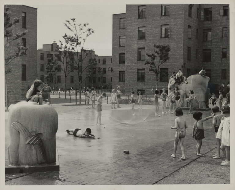 A black and white image from the 1940s shows a courtyard surrounded by brick buildings. In the courtyard, children in swimsuits play in water spraying out from the concrete and climb on stone animal statues surrounding the fountain.