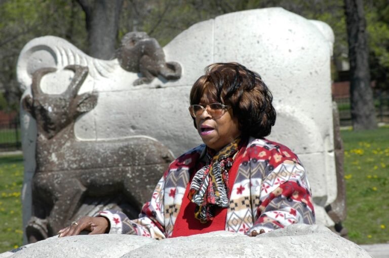 A Black woman in a bright red sweater and sunglasses stands amidst two large stone animal sculptures. She rests her hand on the sculpture in front of her and speaks with passion.