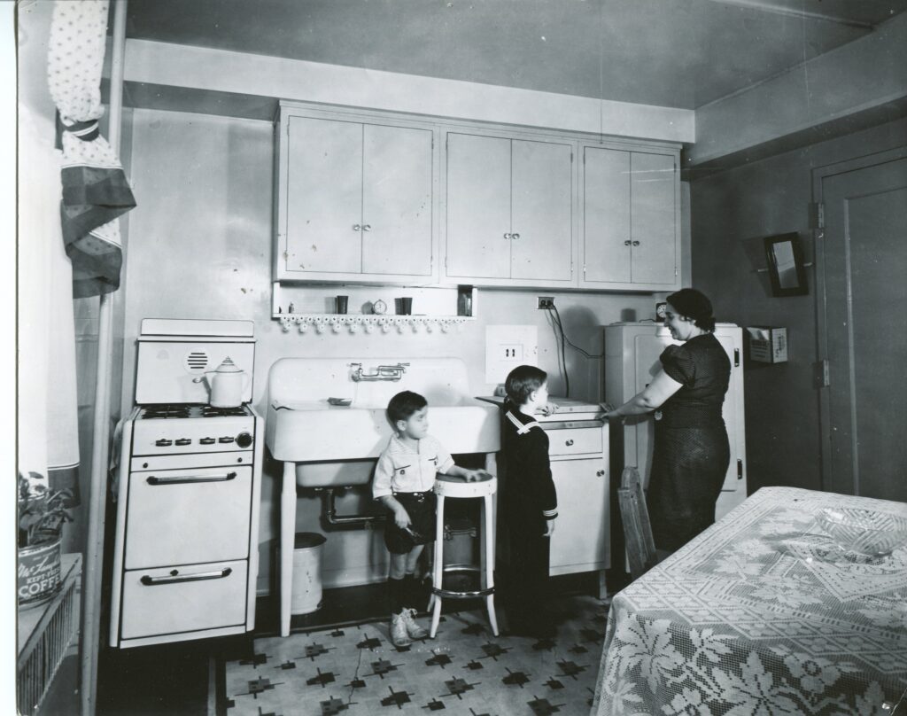 Black and white image of a 1940s Jane Addams Homes kitchen. A mother stands at the counter with her two sons.