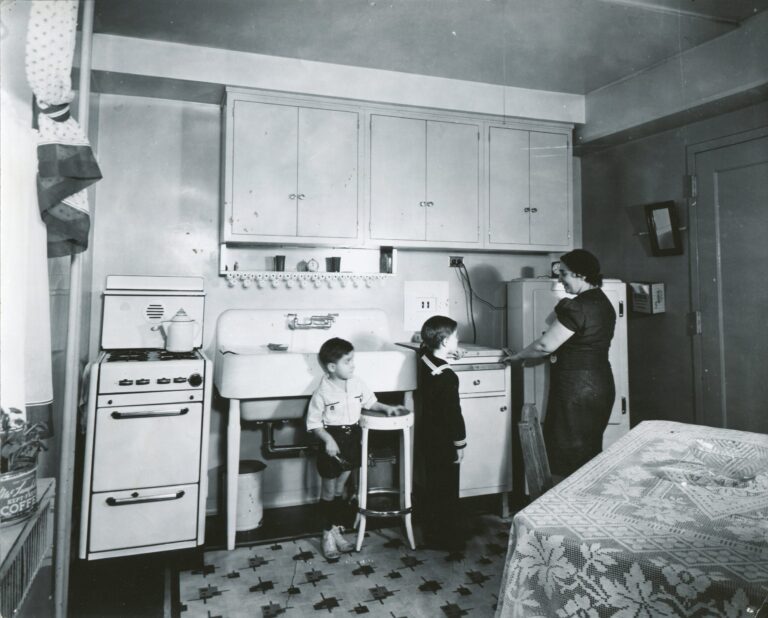 Black and white image of a 1940s Jane Addams Homes kitchen. A mother stands at the counter with her two sons.