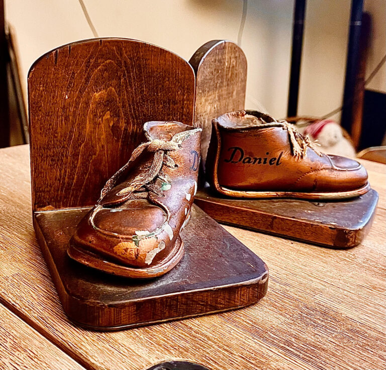 Two wooden bookends, each with a copper baby shoe inscribed with the name “Daniel,” sit on a wooden table.