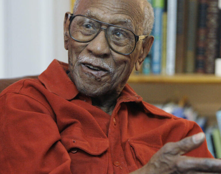 A close up of an older Black man wearing a red shirt and glasses gestures and speaks to someone off camera.