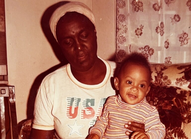 A Black person sits on a couch and holds a baby on her lap