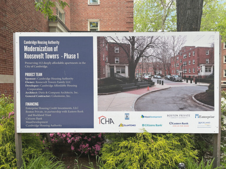 In front of a brick building, a large sign depicts an idyllic street with brick buildings. It reads “Cambridge Housing Authority. Modernization of Roosevelt Towers-Phase 1. Preserving 112 deeply affordable apartments in the City of Cambridge.'' Underneath, it lists project partners and investors.