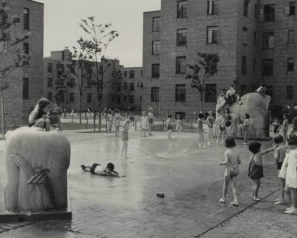 A black and white image from the 1940s shows a courtyard surrounded by brick buildings. In the courtyard, children in swimsuits play in water spraying out from the concrete and climb on stone animal statues surrounding the fountain.