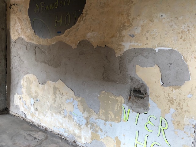 A concrete wall with peeling layers of tan and blue paint. Graffiti in the top left corner says “Abandon Hope.” In the bottom left corner, graffiti that once said “ENTER HERE” is peeling off and obstructed so it only reads “NTER HE.”