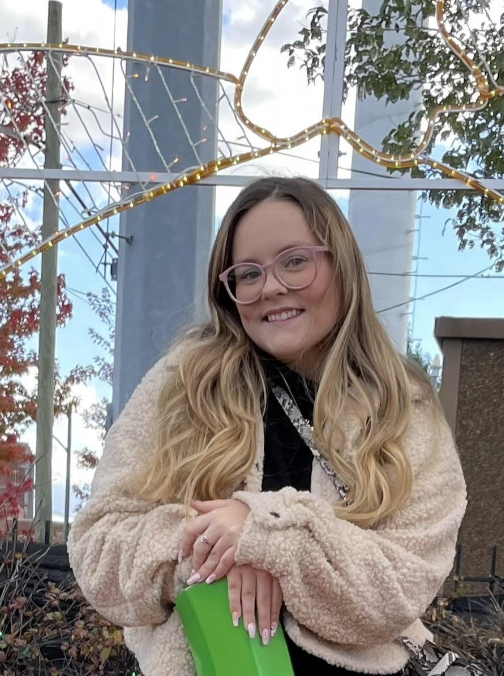 A photo of Alexyss Wofford. Alexyss, a white woman, has long blonde hair and is wearing pink glasses. She has a wooly jacket on and is carrying a green folder. She is smiling slightly. Behind her, there are Christmas lights and some out of season bushes and a tree with red leaves to her right, and a tree with green leaves to her left.