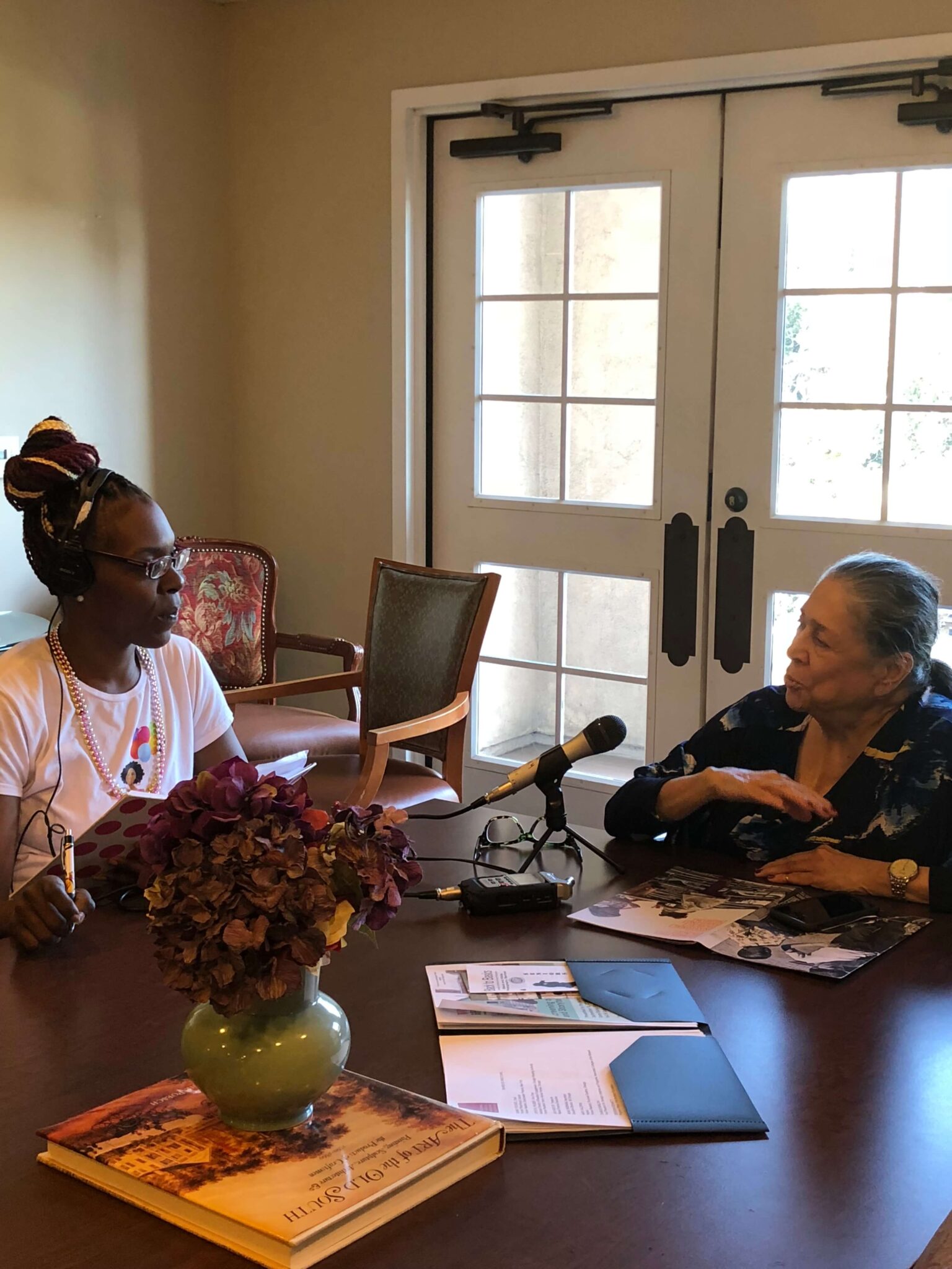 Oral History Collective member Ashley Jefferson (left) interviews an elder, Liz Torres (right). Ashley is wearing headphones and listening attentively as Ms. Torres gestures while telling a story into the recording equipment.