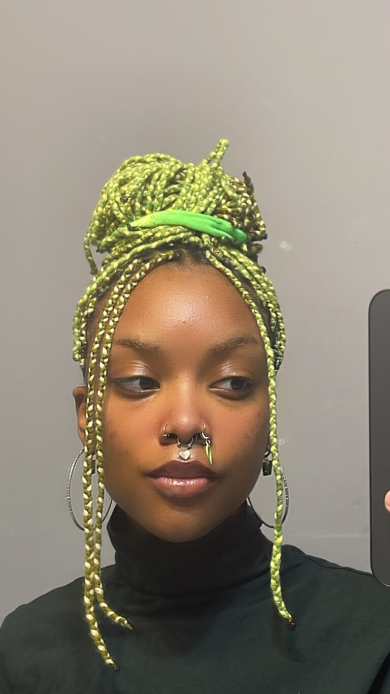 A Black person's mirror selfie. They have green, braided hair, big hooped earrings, and a nose piercing.