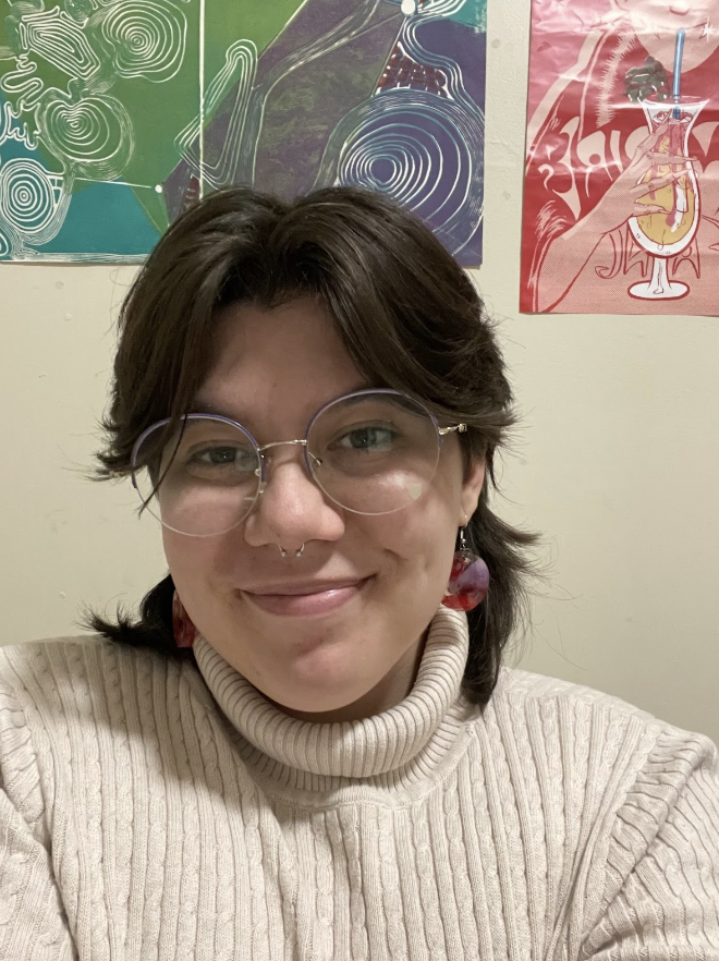 A photo of Sophia Gallo. Sophia, a light-skinned nonbinary person, is wearing thin, silver glasses and two purple and red earrings. Sophia has a nose ring and choppy brown hair that comes to their shoulders. There are two 70s inspired art prints behind her.