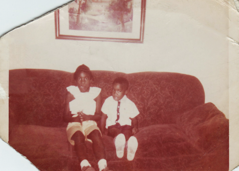 Vintage square photo of two Black children sitting on a couch with a frame hanging on the wall above them.