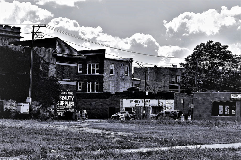 A black and white photo of a neighborhood on the West Side of Chicago. It includes a large vacant lot in the foreground, a couple of neighborhood stores, and housing in the background.