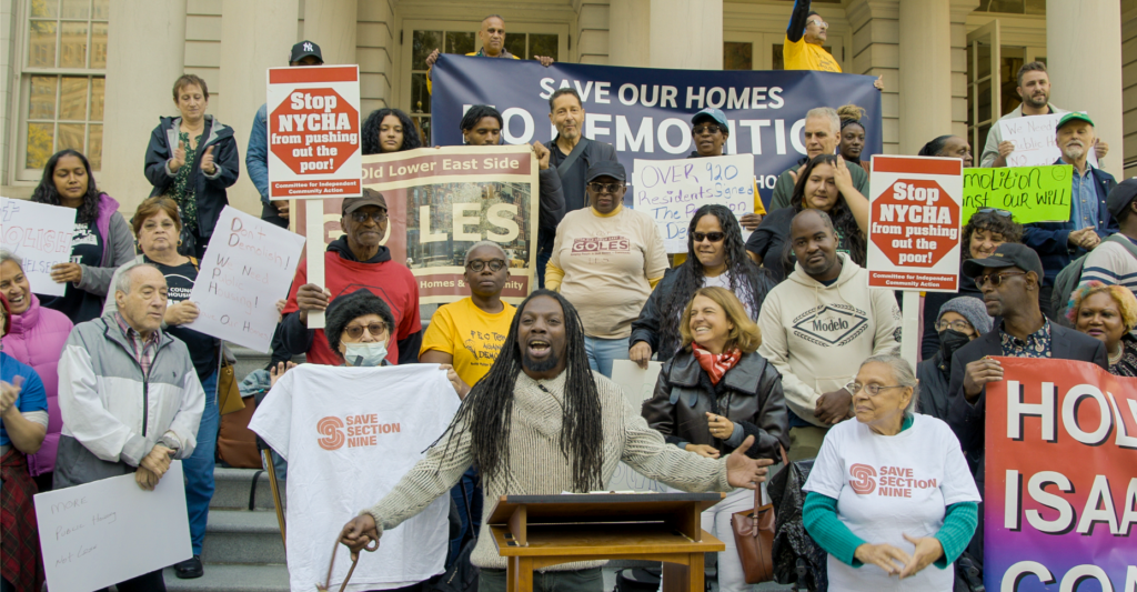 At a rally, a person with long dreadlocks speaks at a podium with a gathering of people behind him. They hold banners, signs, and t-shirts with messages like “Save Section Nine” and “Save our homes.”