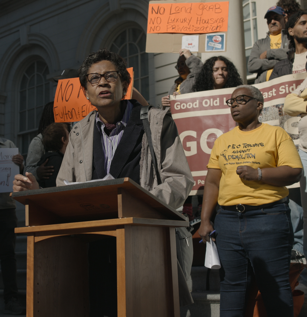 A speaker at a podium with protestors visible in the background