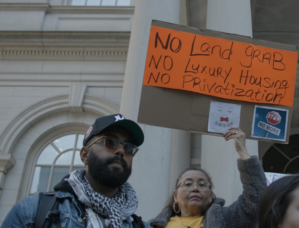 A protestor holds up a handwritten cardboard sign that says "No land grab, no luxury housing, no privatization"