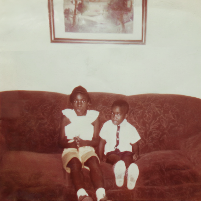 Vintage photo of two Black children sitting on a couch with a frame hanging on the wall above them.