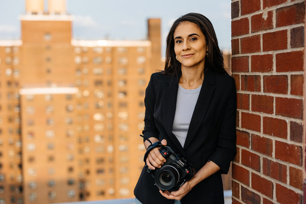 A woman with long dark hair holds a camera and leans against a brick wall on the roof of a building. There is an out-of-focus high-rise in the background.