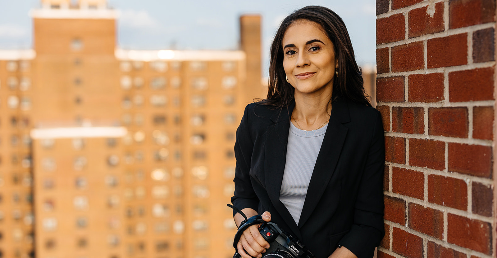 A person in a black blazer and long dark hair holds a camera, leaning against a brick wall with an out-of-focus high-rise in the background.