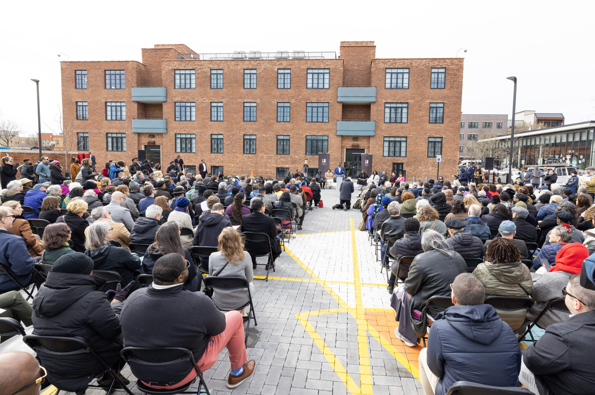 View from behind a seated crowd, as a person speaks at the podium in front of the National Public Housing Museum.