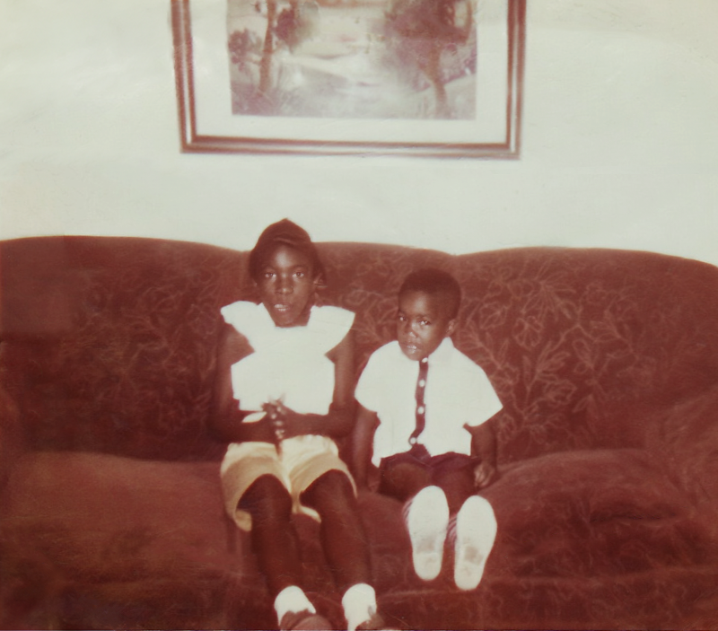 A vintage photo of two Black children sitting on a couch with a frame hanging on the wall above them.
