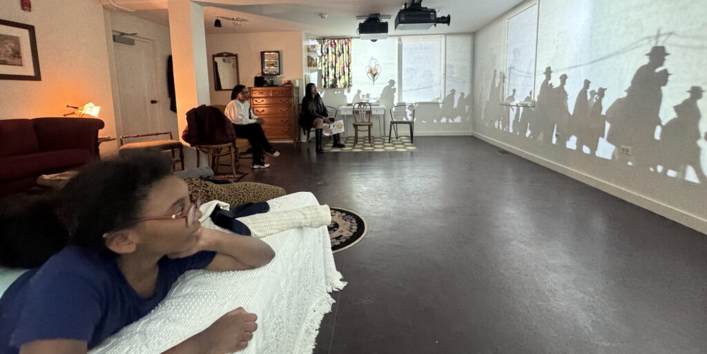 Silhouetted figures are projected on a gallery wall while guests watch from chairs and a bed