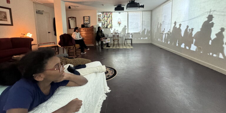 Silhouetted figures are projected on a gallery wall while guests watch from chairs and a bed