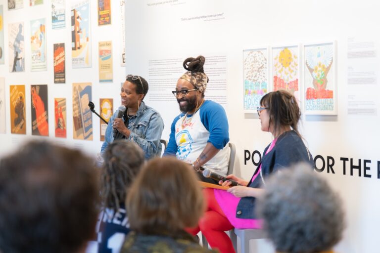 Three smiling presenters sit in front of an audience, with an installation of posters in the background