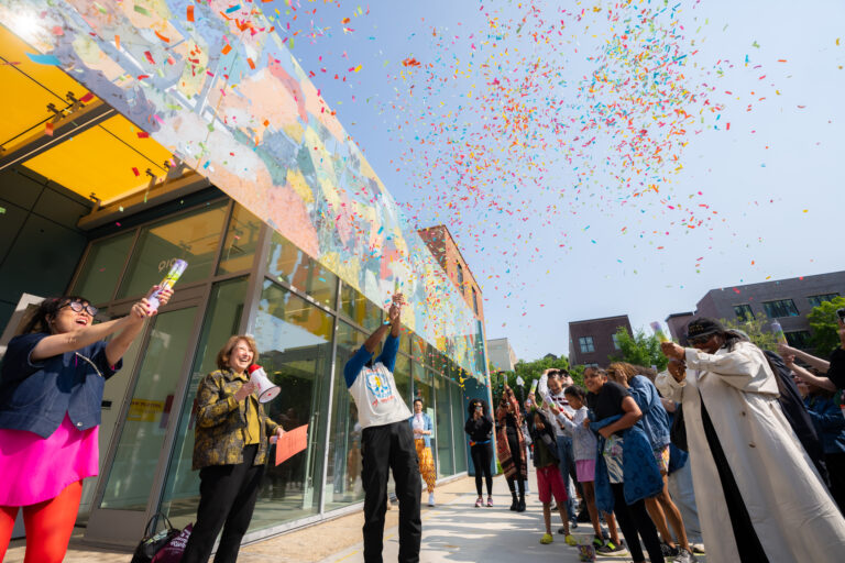 A crowd of people blast confetti into the air on the sidewalk