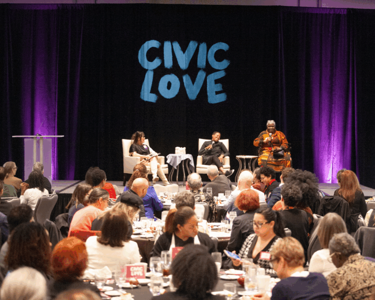 Three presenters on stage in front of a crowd, with the words Civic Love projected in the background