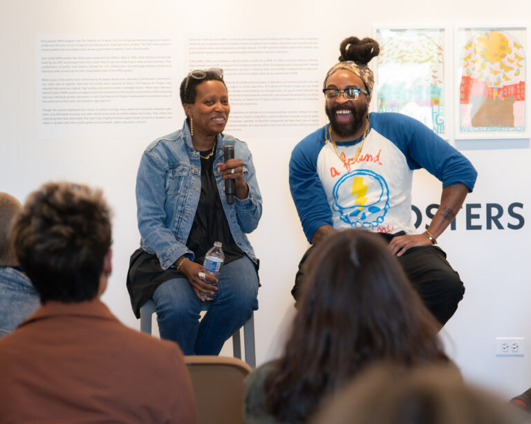 Two people holding mics sit on stools in front of an audience