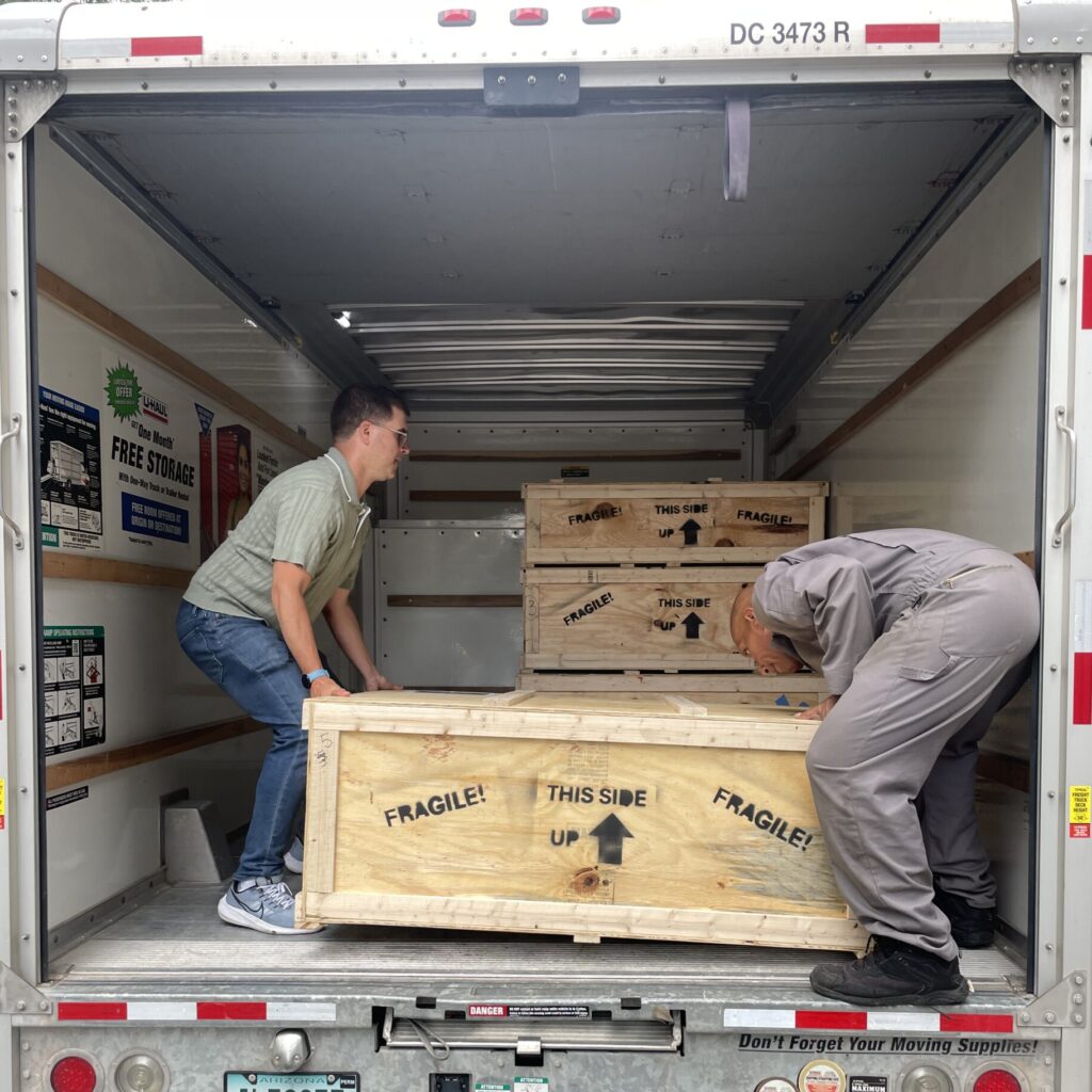 Two people lift a large wooden shipping crate into a truck