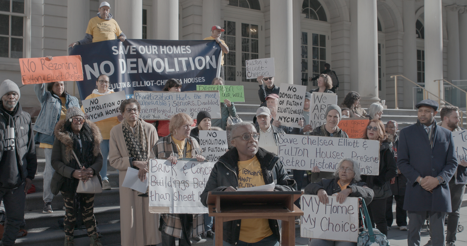 A person speaks at a podium surrounded by a group of protestors holding signs like Save our homes, no demolition.