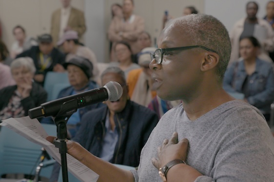 Film still of Renee Keitt speaking at a microphone