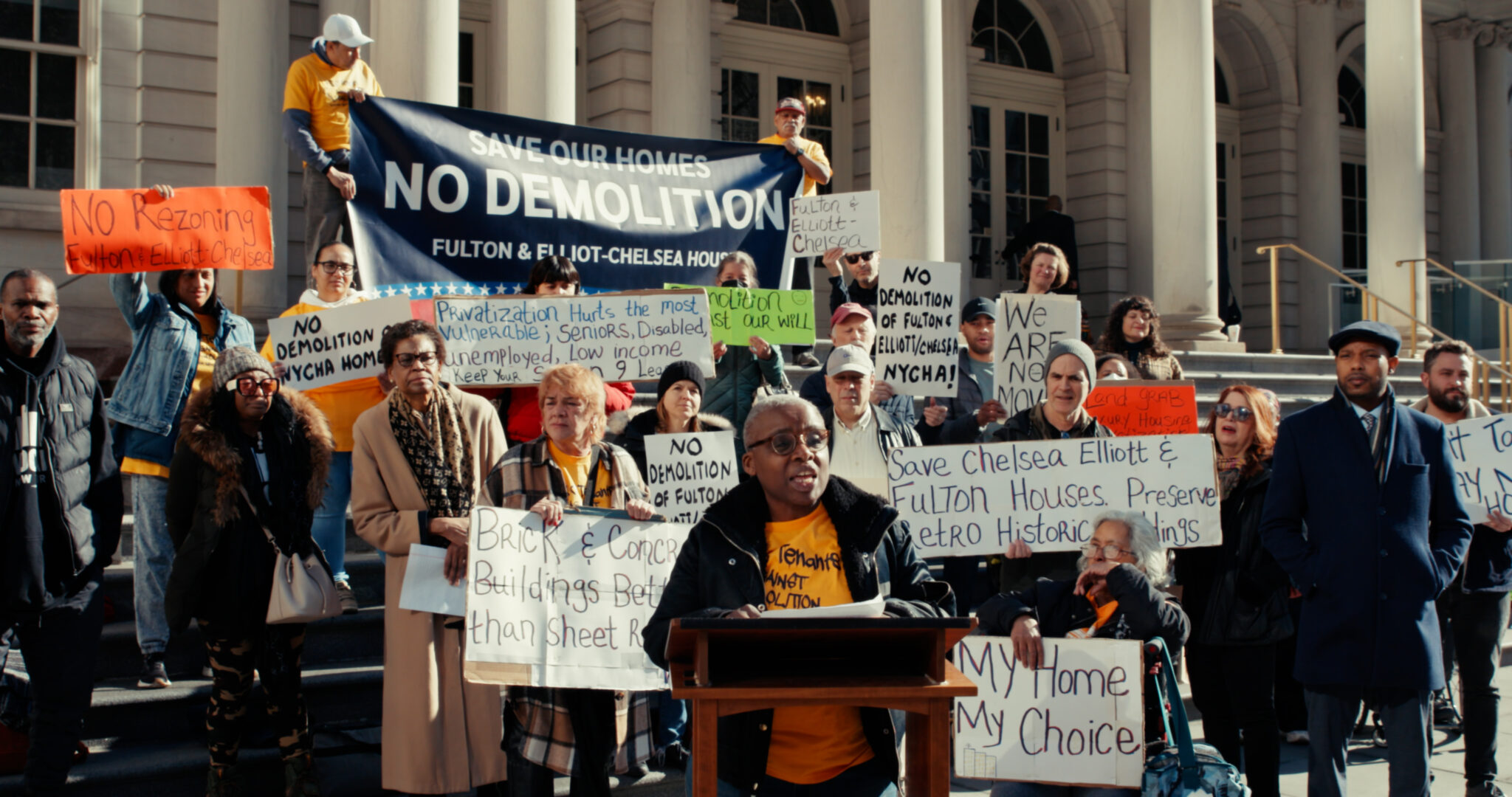 A person speaks at a podium surrounded by a group of protestors holding signs like Save our homes, no demolition.