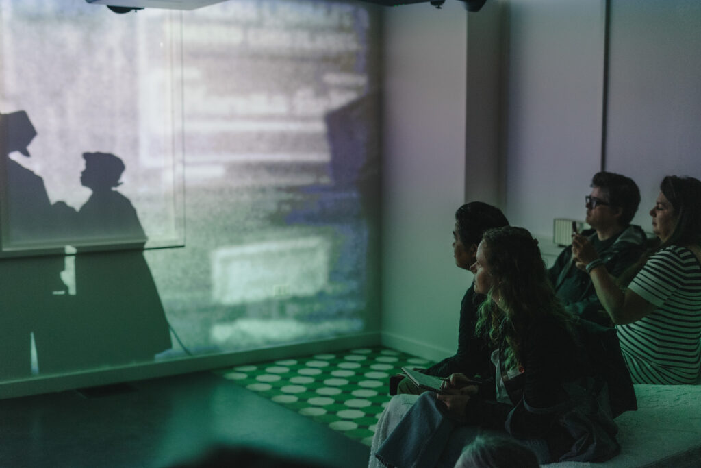 In a dark room, people sit on a bed and watch a projection of silhouette figures on a wall