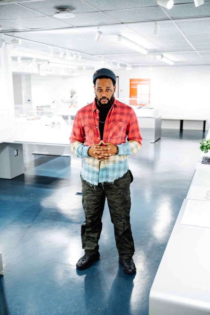 Standing portrait of Open Mike Eagle, in a red-yellow-blue flannel shirt, camo pants, and knit beanie