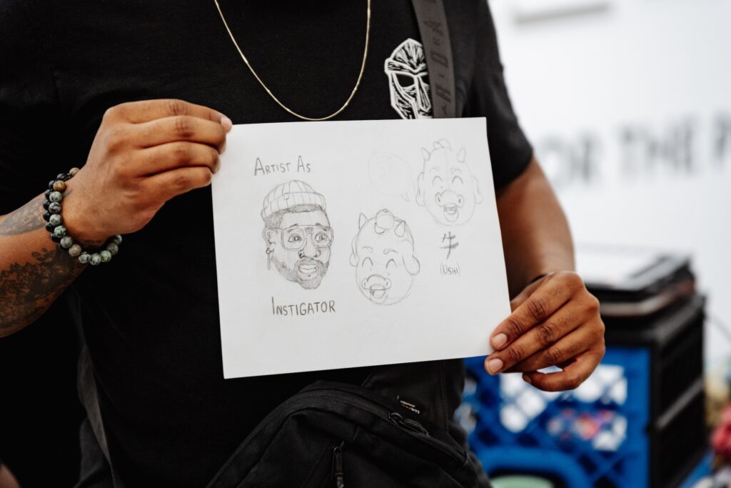A person holds up a pencil sketch of Open Mike Eagle with the words Artist as Instigator