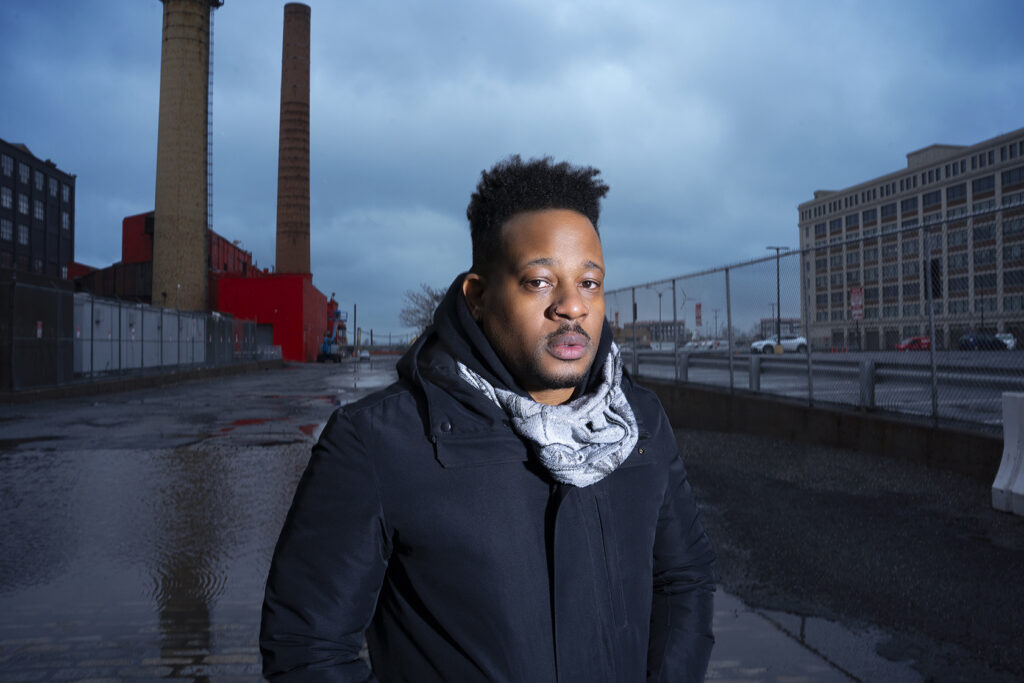 Portrait of Open Mike Eagle standing outside with smokestacks and a chain link fence in the background