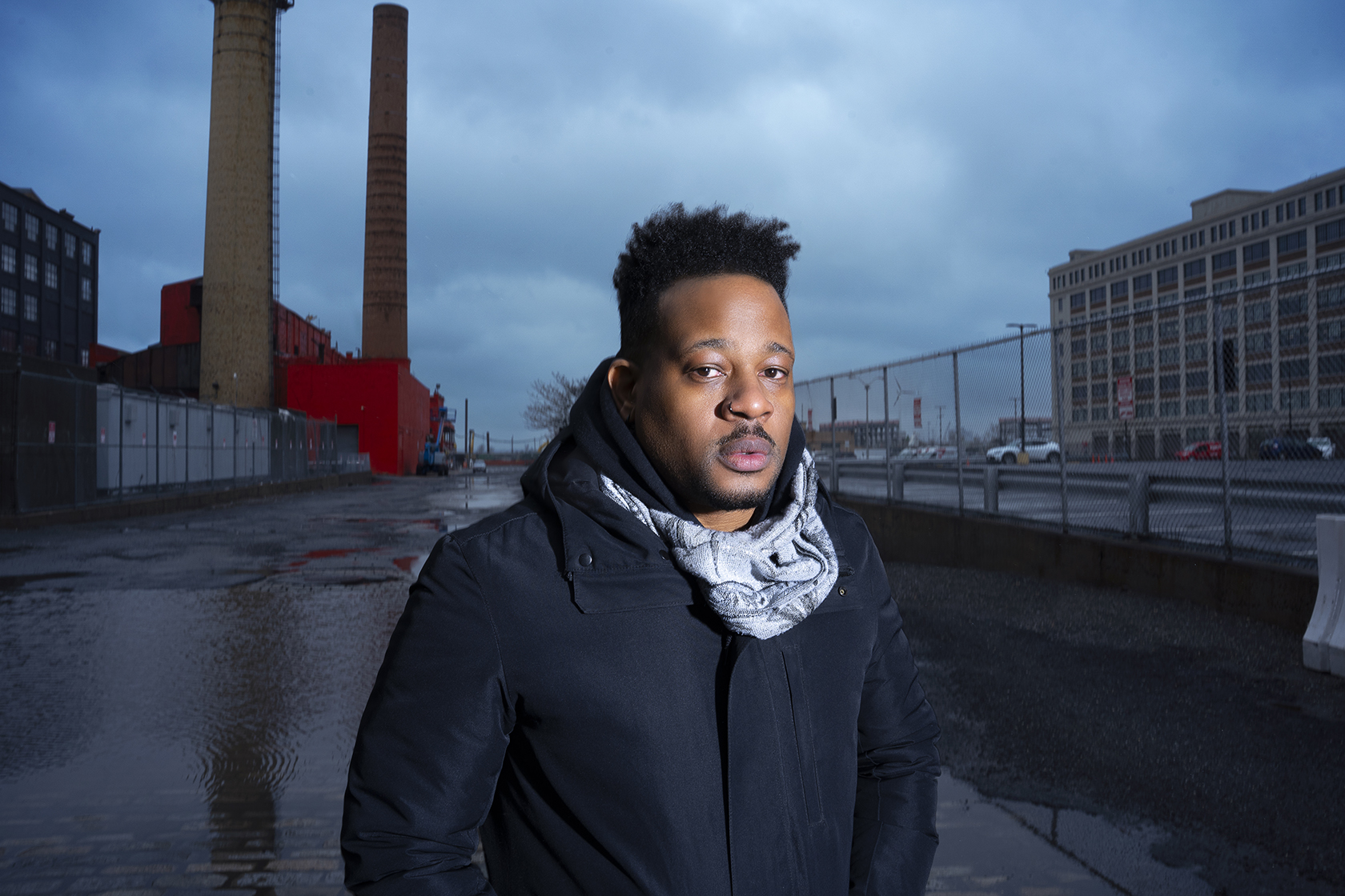 Portrait of Open Mike Eagle standing outside with smokestacks and a chain link fence in the background