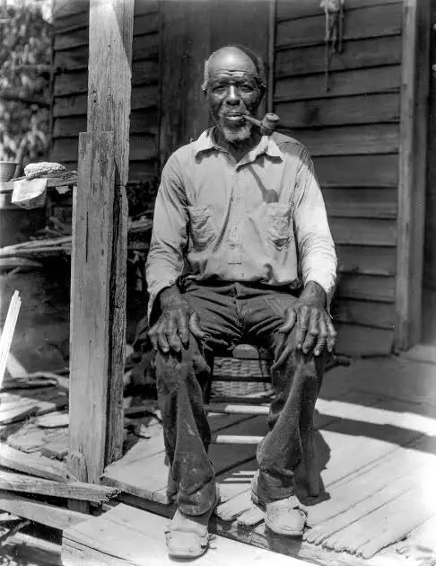 A c. 1930s black and white photo of a Black man sitting on a porch with a pipe in his mouth.
